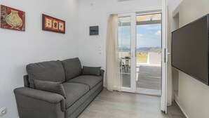 A living room with a grey sofa, a flat-screen TV, and a sliding glass door leading to a balcony with a view of the countryside. - Sunset Village (Milos)