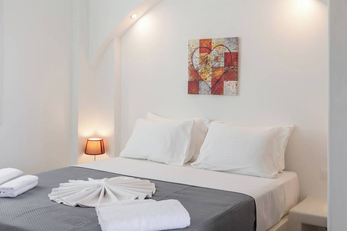 A hotel room with a bed, white pillows, a gray bedspread, a wall-mounted artwork, and a bedside lamp.