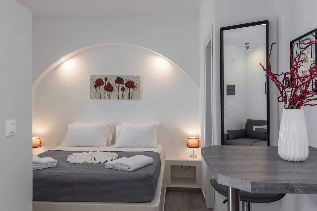 A modern hotel room with a curved ceiling, a bed with white linens, a bedside table with a lamp, a mirror, and a vase with red branches.
