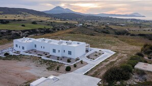 A modern white building with multiple satellite dishes, surrounded by a landscaped garden with small bushes and a paved driveway. - Sunset Village (Milos)