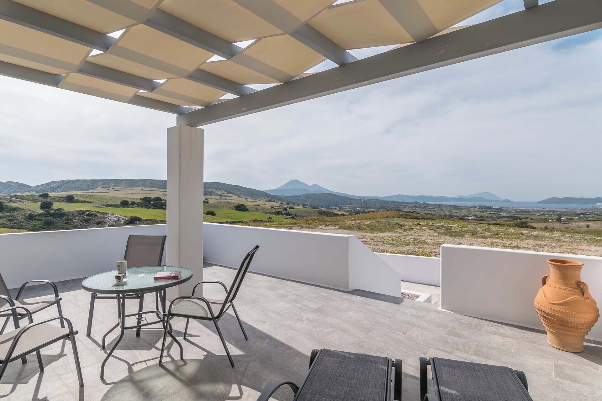 A terrace with a glass table, metal chairs, and a view of a hilly landscape.