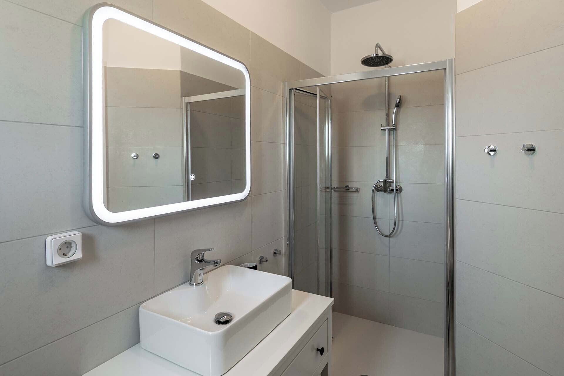 Une salle de bain moderne avec un lavabo blanc, un miroir doté d’un cadre lumineux et une douche à parois de verre.