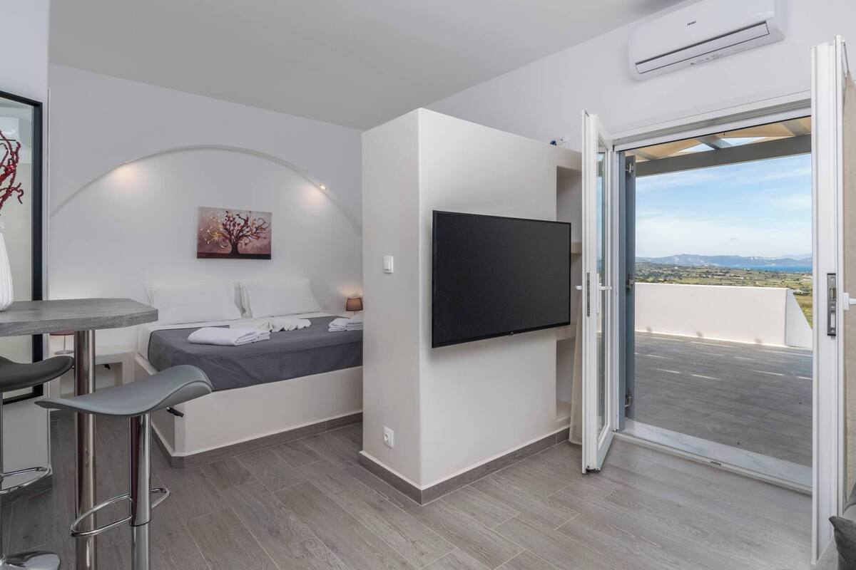 A modern hotel room with a bed, a TV mounted on the wall, a small table with chairs, and a balcony with a view.