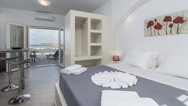 A hotel room with a bed, a desk, and a chair. There is a view of the sea through the open door. - Sunset Village (Milos)