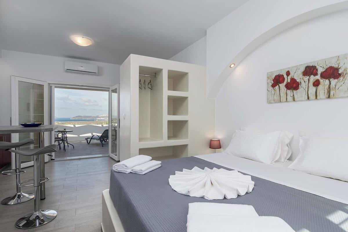 A hotel room with a bed, a desk, and a chair. There is a view of the sea through the open door.
