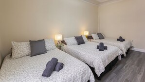 Ein Hotelzimmer mit vier Betten, jedes mit floralem Überwurf und dunklem Kissen. Nachttischlampen und ein kleiner Tisch befinden sich zwischen den Betten.