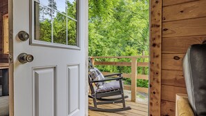 A wooden deck with a rocking chair and a bench, a white door with a window, and a view of green trees.