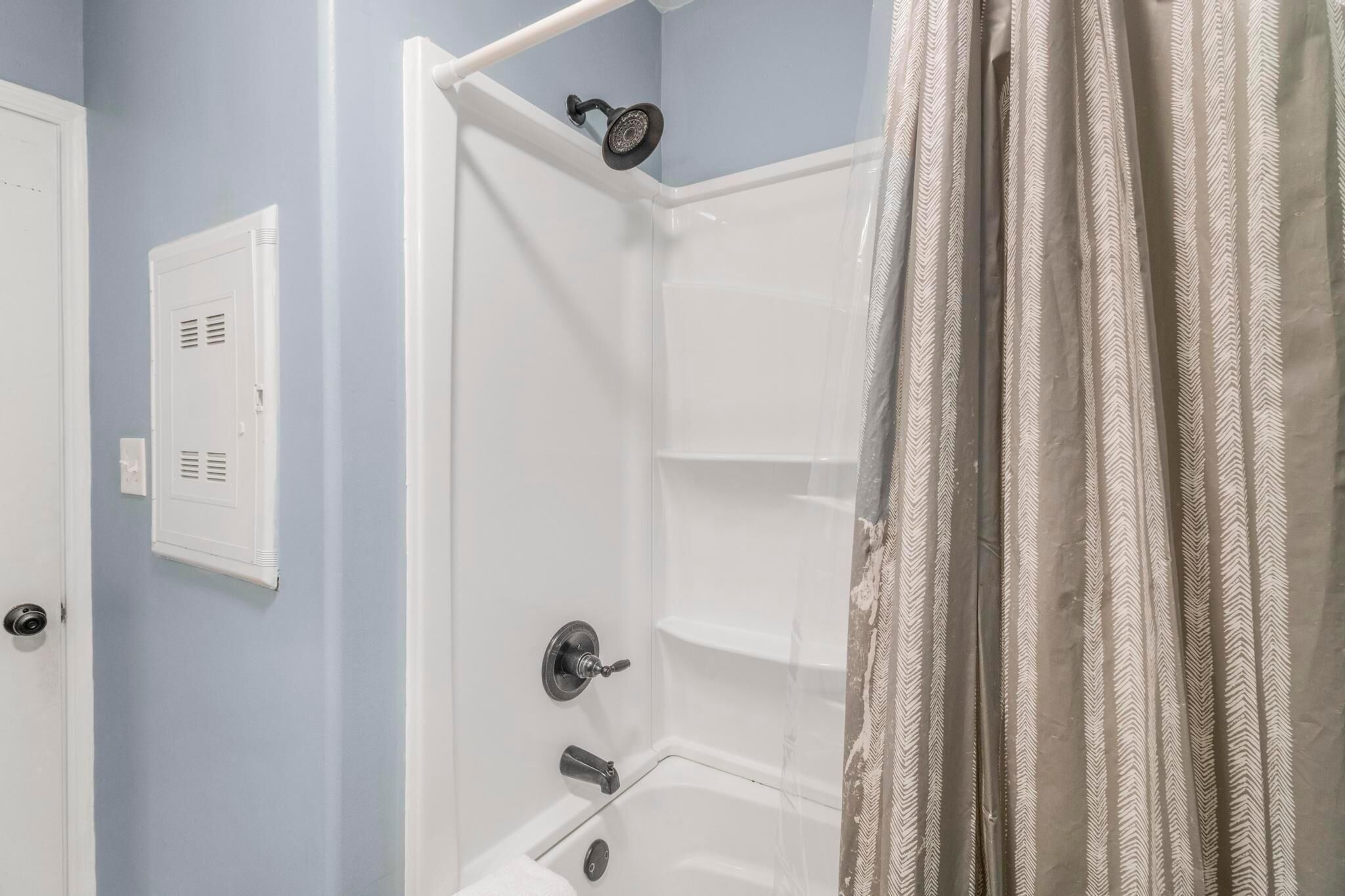 Una ducha con cabezal de ducha de mano, una puerta blanca con rejilla de ventilación y una cortina estampada.