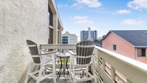 A balcony with two Adirondack chairs and a small table, overlooking a building with a sign that reads "SUNDAL". - The Village 15D (Gulf Shores)