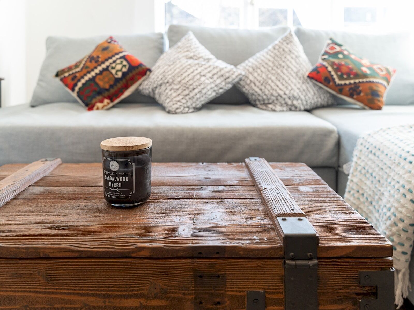 Un coffre en bois rustique, surmonté d’une bougie, est posé sur une table en bois dans un salon chaleureux, où se trouvent un canapé gris et des coussins colorés.