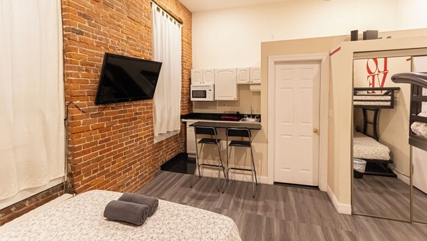 A compact hotel room with a brick wall, a bed with a patterned bedspread, a small kitchenette, and a mirror reflecting a bunk bed. - Cozy STUDIO 1Full/2Twin beds near Empire State Building   (New York)