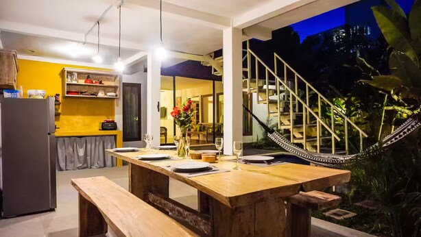 A dining area with a wooden table set for four, a kitchen in the background, and a staircase leading to an upper level.