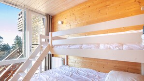 Una cama nido con ropa de cama blanca, cabecera de madera y ventana con vista a árboles.