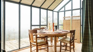 Un invernadero con una mesa redonda dispuesta para comer, rodeada de sillas de madera con cojines.
