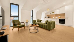 Ein modernes Wohnzimmer mit einem grünen Sofa, einem Holztisch und einem Sessel. Es gibt eine Küchenzeile mit weißen Schränken und einer Holz Arbeitsplatte.