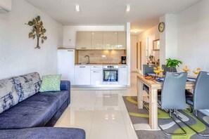 A modern kitchen with white cabinets, a dining area with a table and chairs, and a living room with a sofa and a decorative wall piece. - Holiday apartment with sea view (Njivice)