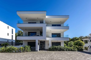 A modern multi-story building with balconies, a paved entrance, and surrounding greenery.
