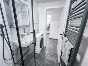 A modern bathroom with a glass shower enclosure, a white sink, a washing machine, and a towel rack.