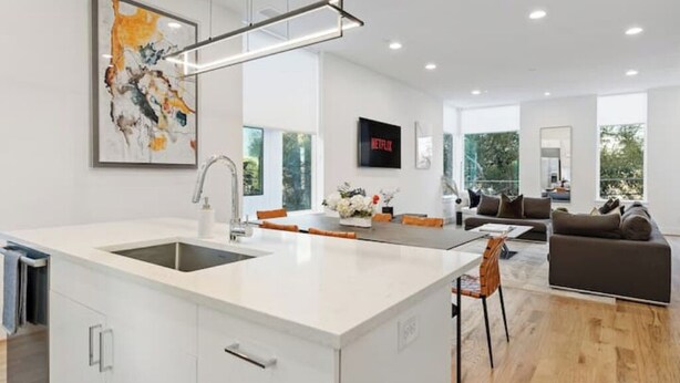 Una cocina moderna con una isla central, una zona de comedor con mesa y sillas, una sala de estar con sofá y un amplio ventanal que proporciona luz natural.