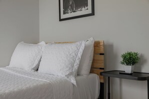 A neatly made bed with white bedding, a wooden headboard, a framed picture on the wall, and a small potted plant on a side table. - Modern & Spacious~Rooftop View of Downtown Dallas (Dallas)