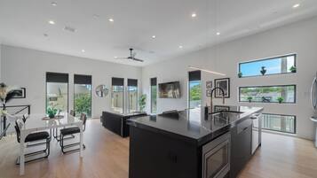 Uma cozinha moderna com ilha central, grandes janelas e área de jantar com mesa e cadeiras brancas.