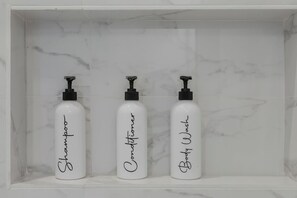 Three white bottles with black pumps labeled 'Shampoo,' 'Conditioner,' and 'Body Wash' in a white marble shelf. - Modern & Spacious~Rooftop View of Downtown Dallas (Dallas)