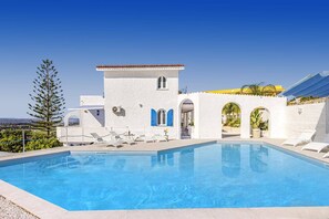 Un área de piscina con una piscina azul cristalina, paredes blancas, contraventanas azules y vistas al paisaje.