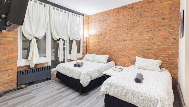 Una habitación con dos camas, una pared de ladrillo, una ventana con cortinas y un televisor de pantalla plana montado en la pared.
