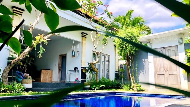 Uma piscina com superfície de água azul cristalina, rodeada por vegetação exuberante e um edifício branco com porta e janelas.