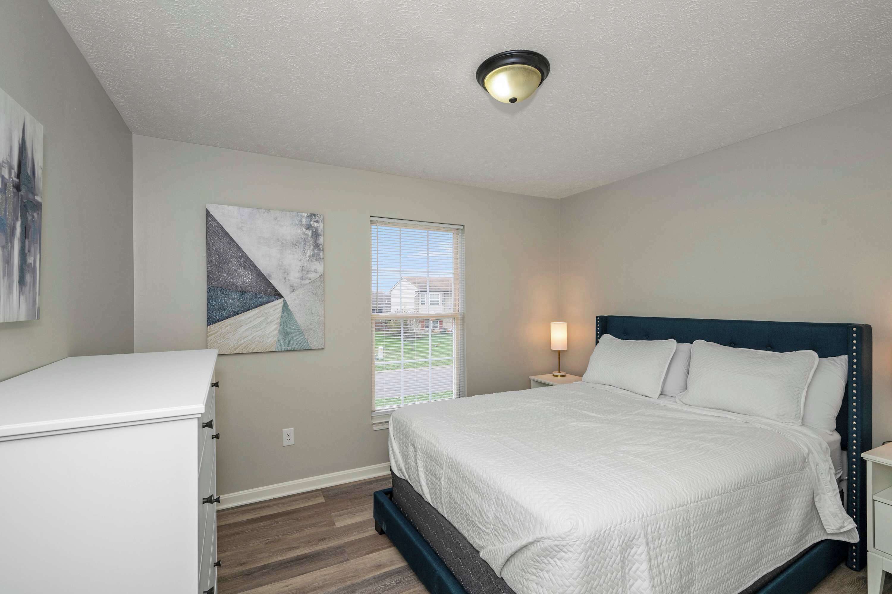 A bedroom with a bed, a nightstand, a dresser, a window, and a ceiling light.