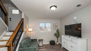 A living room with a staircase, a television on a white cabinet, a green sofa, a small table, and a window with blinds. - The Restful Ark (Crittenden)