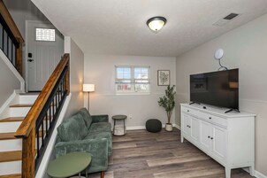 Una sala de estar con una escalera, un televisor sobre un mueble blanco, un sofá verde, una mesita y una ventana con persianas.