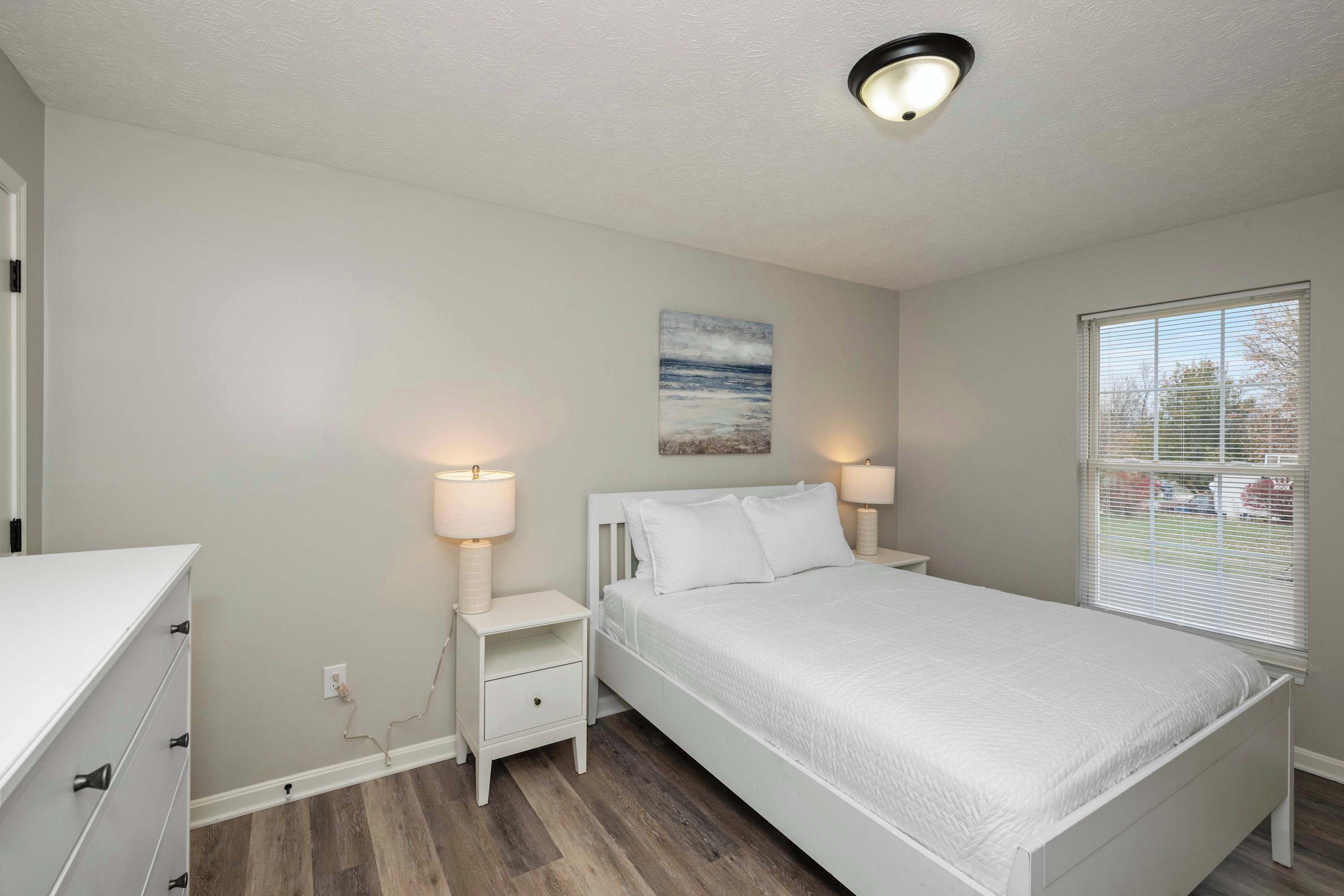 A bedroom with a bed, bedside lamps, a nightstand, a dresser, and a window with blinds.