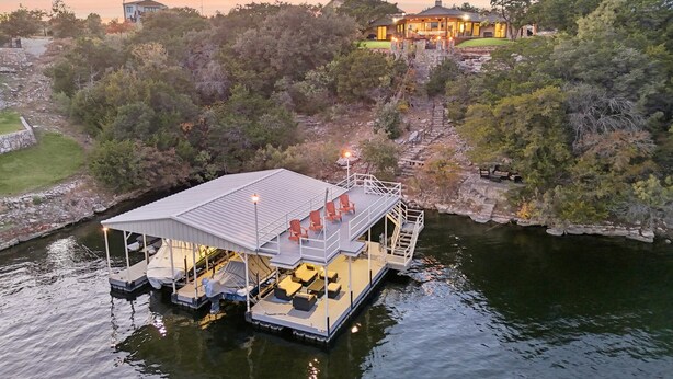 Ein schwimmendes Haus mit überdachter Terrasse und Außenmöbeln, umgeben von Bäumen und einem Gewässer.