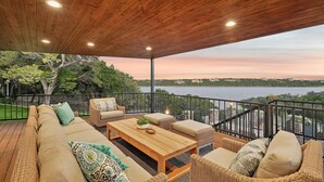 Eine Holzterrasse mit einer Couch, Sesseln und einem Tisch, mit Blick auf ein Gewässer und Grünflächen.