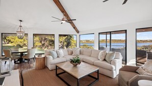 Ein geräumiges Wohnzimmer mit einer großen Eckcouch, einem Couchtisch und einem Essbereich mit Stühlen. Große Fenster bieten Blick auf einen See und Bäume.
