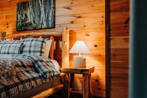 A wooden cabin room with a bed, plaid bedding, a painting of a forest, and a table with a lamp.