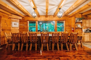 A wooden cabin interior with a dining table surrounded by chairs, a kitchen area, and large windows with a view of greenery.