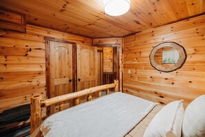 A wooden cabin interior with a bed, wooden walls, a circular mirror, and a door.