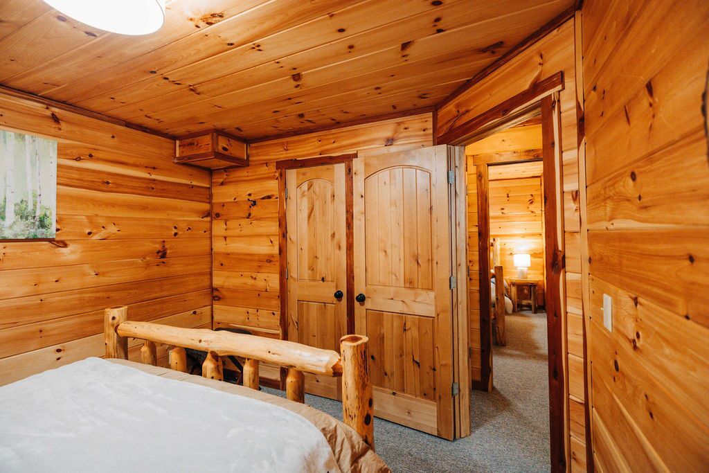 A wooden cabin interior with a bed, wooden walls, and a door leading to another room.