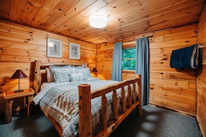 Une chambre dans une cabane en bois, avec un lit, une table de chevet, une lampe, une fenêtre avec des rideaux et une serviette accrochée au mur.