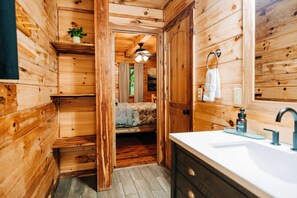 L’intérieur d’un chalet en bois comprend un lit, une salle de bain avec un lavabo et un miroir, ainsi qu’un ventilateur de plafond.