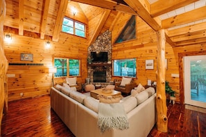 A log cabin interior with a stone fireplace, wooden walls, a large window, and a cozy seating area.