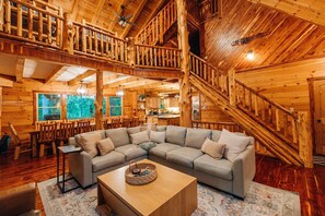 L’intérieur d’une cabane en rondins, avec un grand canapé d’angle en forme de L, un coin repas et un escalier.