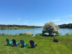 A grassy area with four Adirondack chairs facing a lake, surrounded by trees and a clear sky.