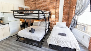 A dormitory room with bunk beds, a kitchenette, and a brick wall.