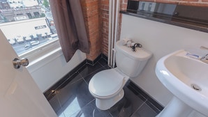 A small bathroom with a toilet, sink, and window overlooking a street.