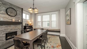 Un espace repas comprenant une longue table, des chaises, une cheminée, une horloge et une fenêtre avec des stores.