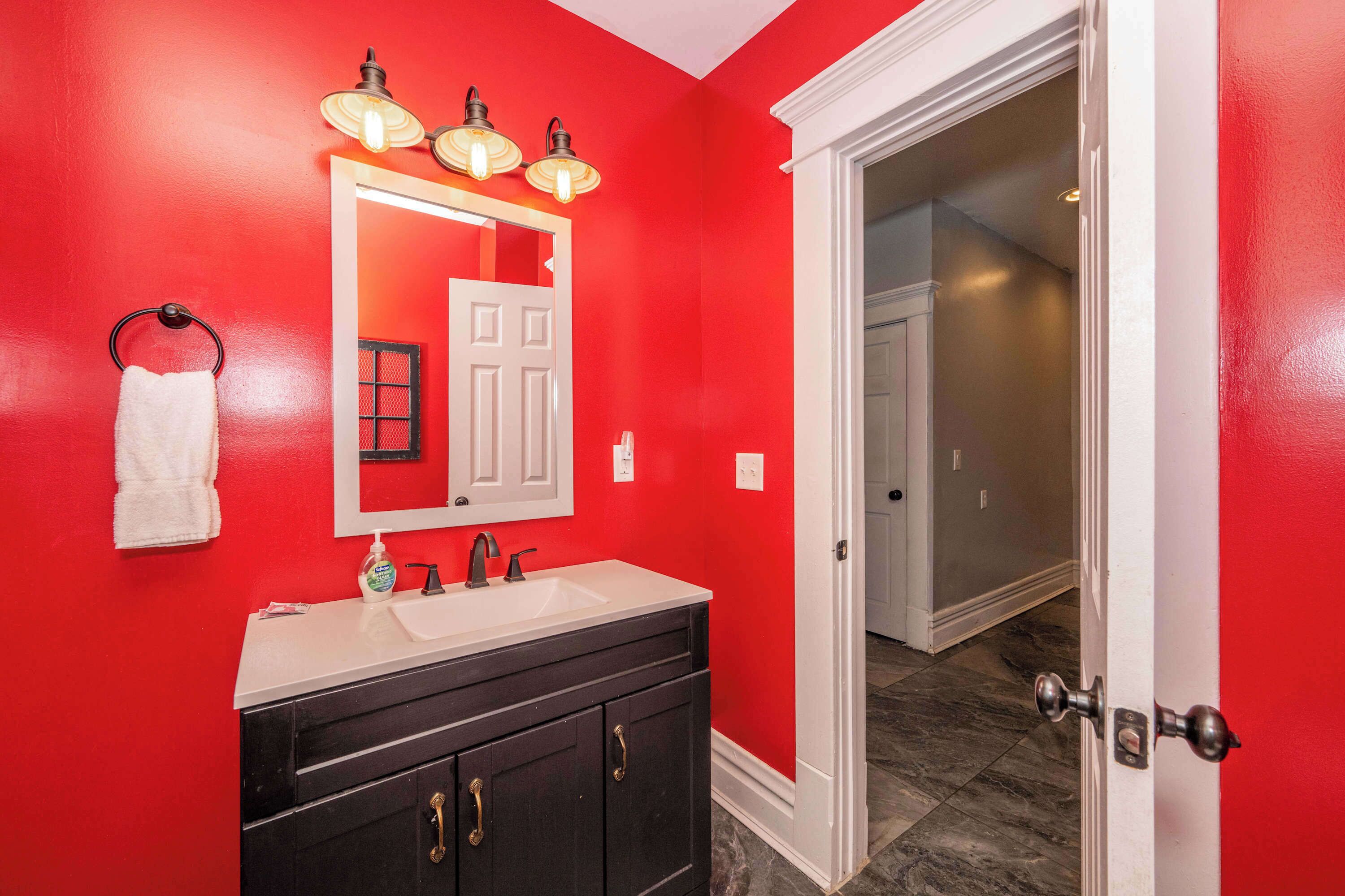 Une salle de bain avec un mur rouge, une porte blanche et un meuble-lavabo noir doté d’un miroir et d’éclairages.