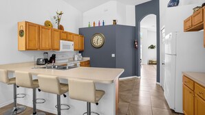 Une cuisine avec des meubles en bois, un micro-ondes blanc, une horloge et un coin bar avec des tabourets.